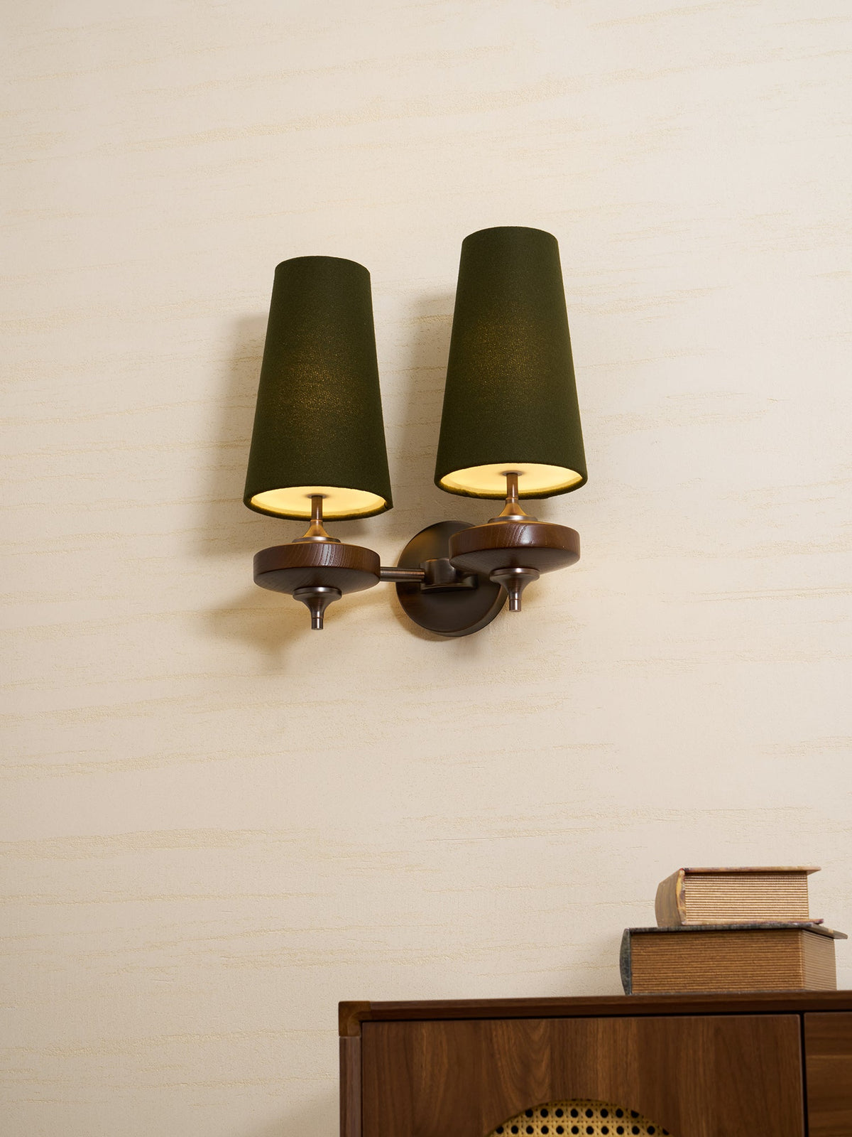 Lit green shades and brass-toned arms on a Clérachic wall light, mounted on a pale wall over a walnut cabinet and a pile of books.