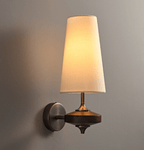 A single cream shade lit warm above a dark wood disc and round bronze backplate on a Clérachic wall sconce, against a grey wall.