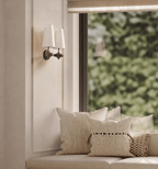 A pair of white shades on a Clérachic wall sconce beside a window, above a bench piled with cream cushions.