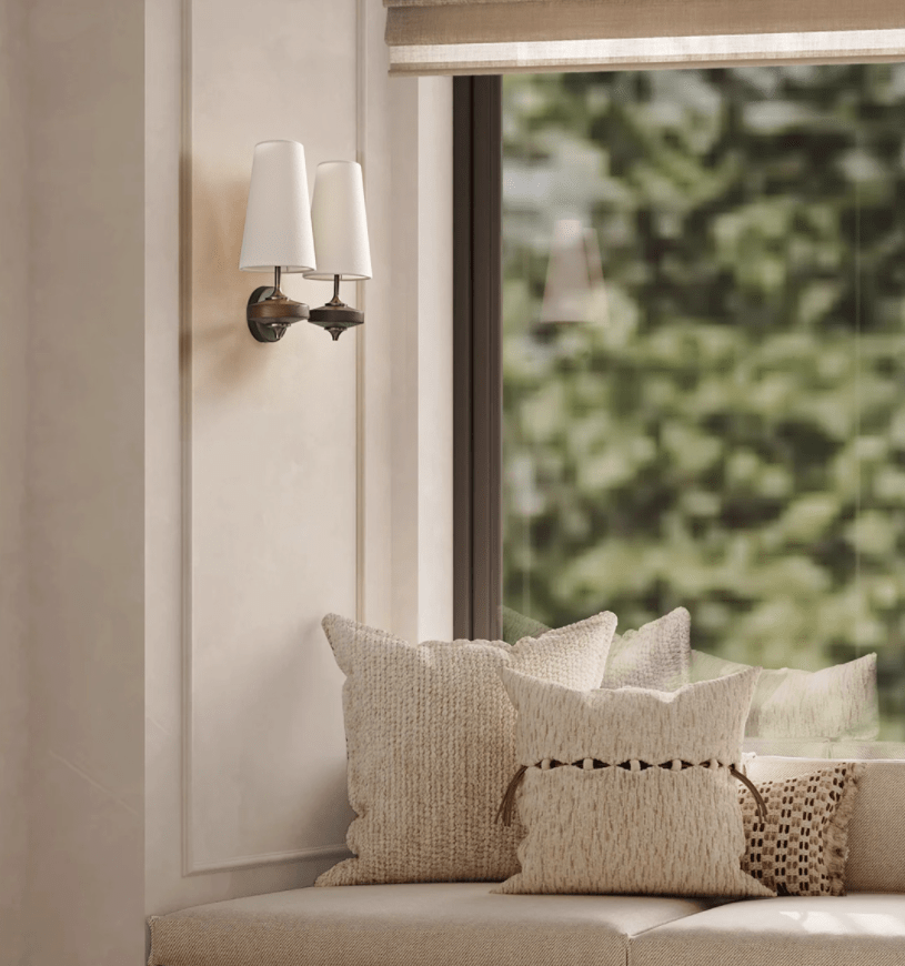A pair of white shades on a Clérachic wall sconce beside a window, above a bench piled with cream cushions.