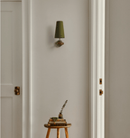 A lone green-shaded Clérachic wall light on a white panelled wall between two doors, above a small stool with books.