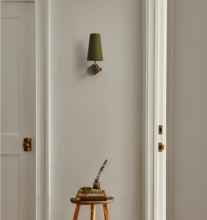 A lone green-shaded Clérachic wall light on a white panelled wall between two doors, above a small stool with books.