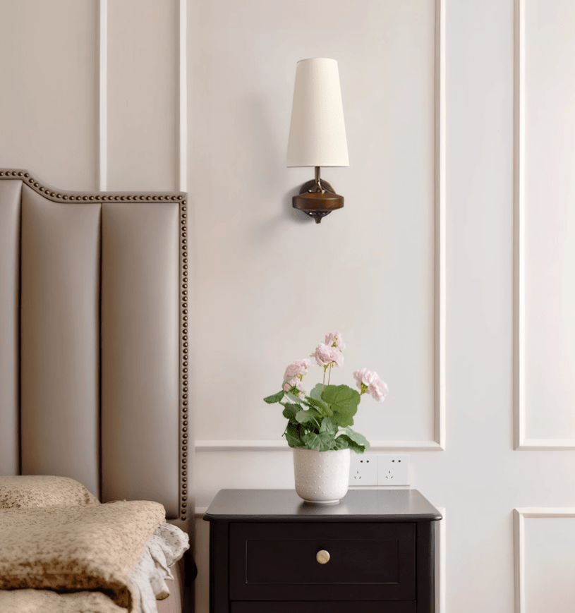 A single white-shaded Clérachic wall sconce beside a padded headboard, above a dark bedside table with pink flowers.
