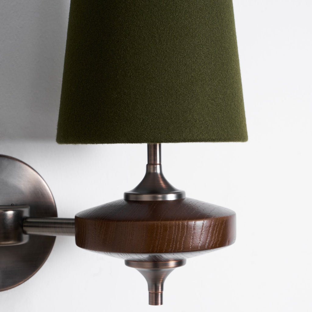 Close view of a green felt-like shade, bronze stem and grained walnut disc on this Clérachic wall sconce, on white.