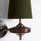 Close view of a green felt-like shade, bronze stem and grained walnut disc on this Clérachic wall sconce, on white.