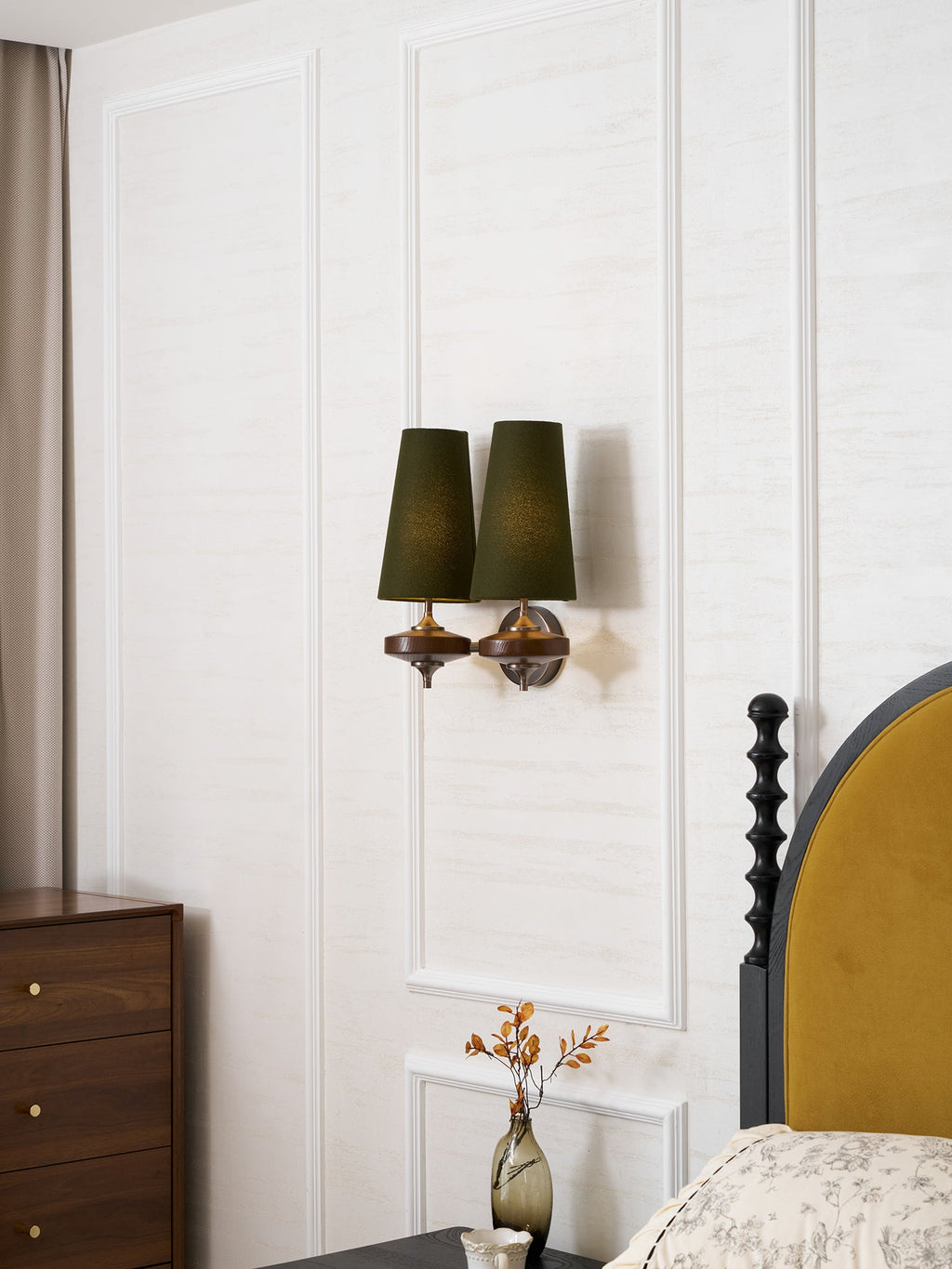 A Clérachic twin green-shaded wall sconce on a white panelled wall, above a walnut chest of drawers and a bed with a mustard headboard.