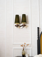 Lit from below, a Clérachic two-light sconce in green hangs on a white panelled wall beside a mustard-velvet bedhead.
