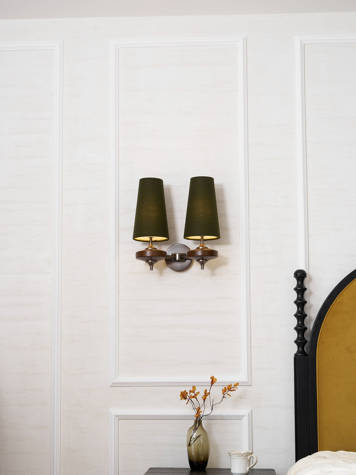 A small Clérachic twin wall light with green shades on a white panelled wall, above a vase of autumn stems.