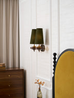 Angled view of a Clérachic green twin sconce on a panelled wall, with a taupe curtain and a walnut chest of drawers beside it.