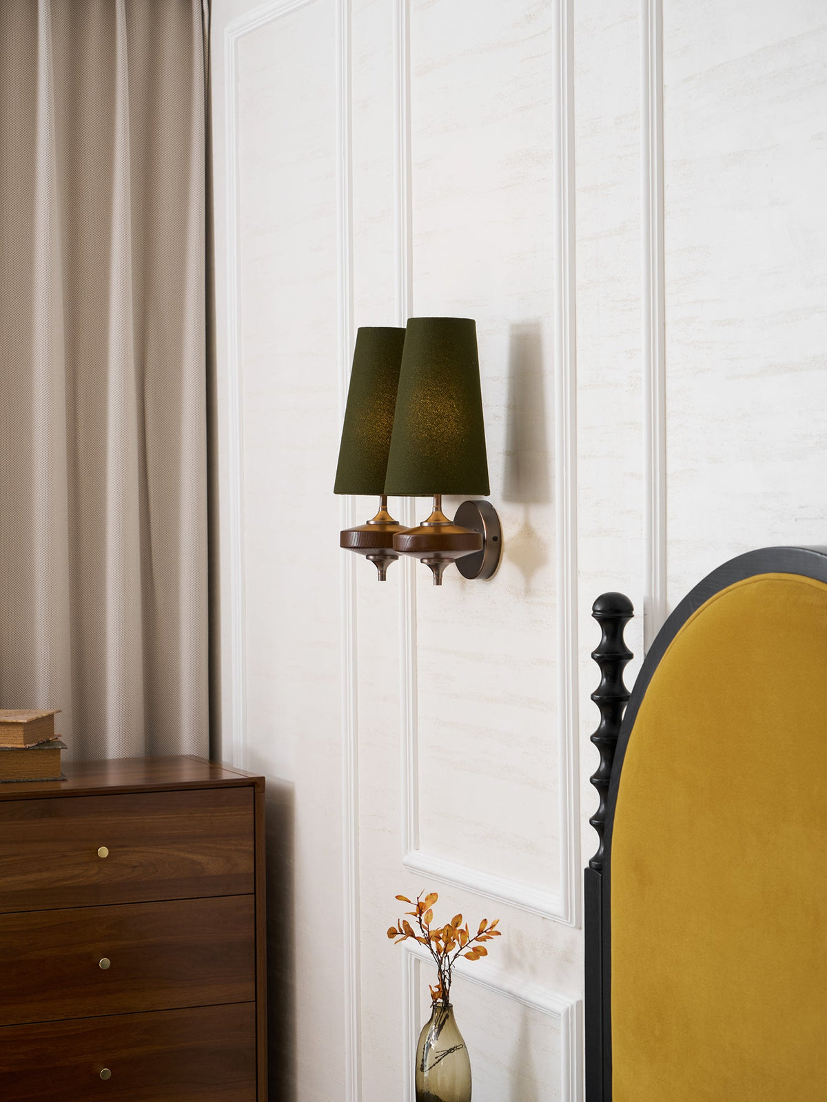 Angled view of a Clérachic green twin sconce on a panelled wall, with a taupe curtain and a walnut chest of drawers beside it.