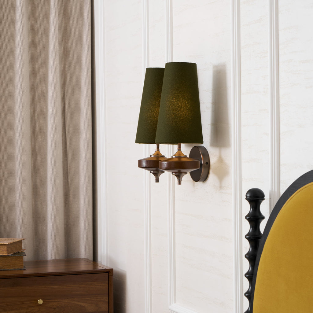 A Clérachic twin sconce with green shades seen from the side, beside a taupe curtain and a mustard bedhead.