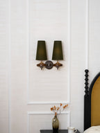 Two green shades on a Clérachic wall sconce, centred in a white wall panel above a bedside table with a glass vase.