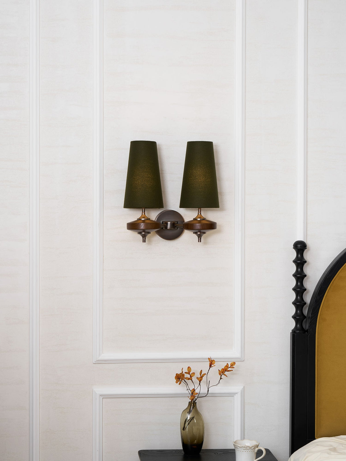 Two green shades on a Clérachic wall sconce, centred in a white wall panel above a bedside table with a glass vase.