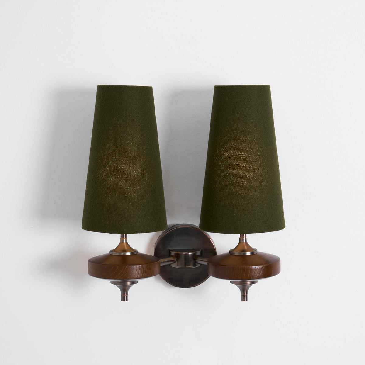 Softly lit green shades on a Clérachic twin wall light, with walnut discs and a bronze link arm on a plain white wall.
