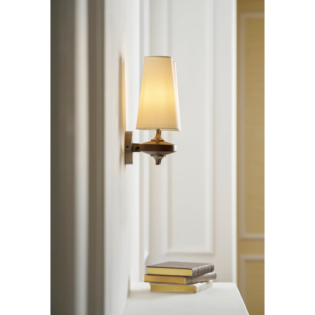 A single cream shade glowing beside a white door on a Clérachic wall sconce, above a stack of books on a ledge.