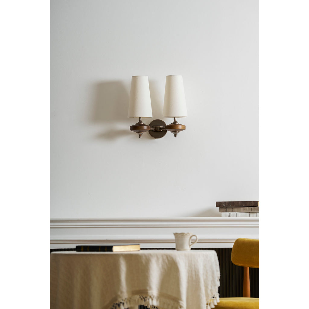 A Clérachic twin wall sconce with cream shades over a white mantel, a small jug and a round table with a linen cloth.