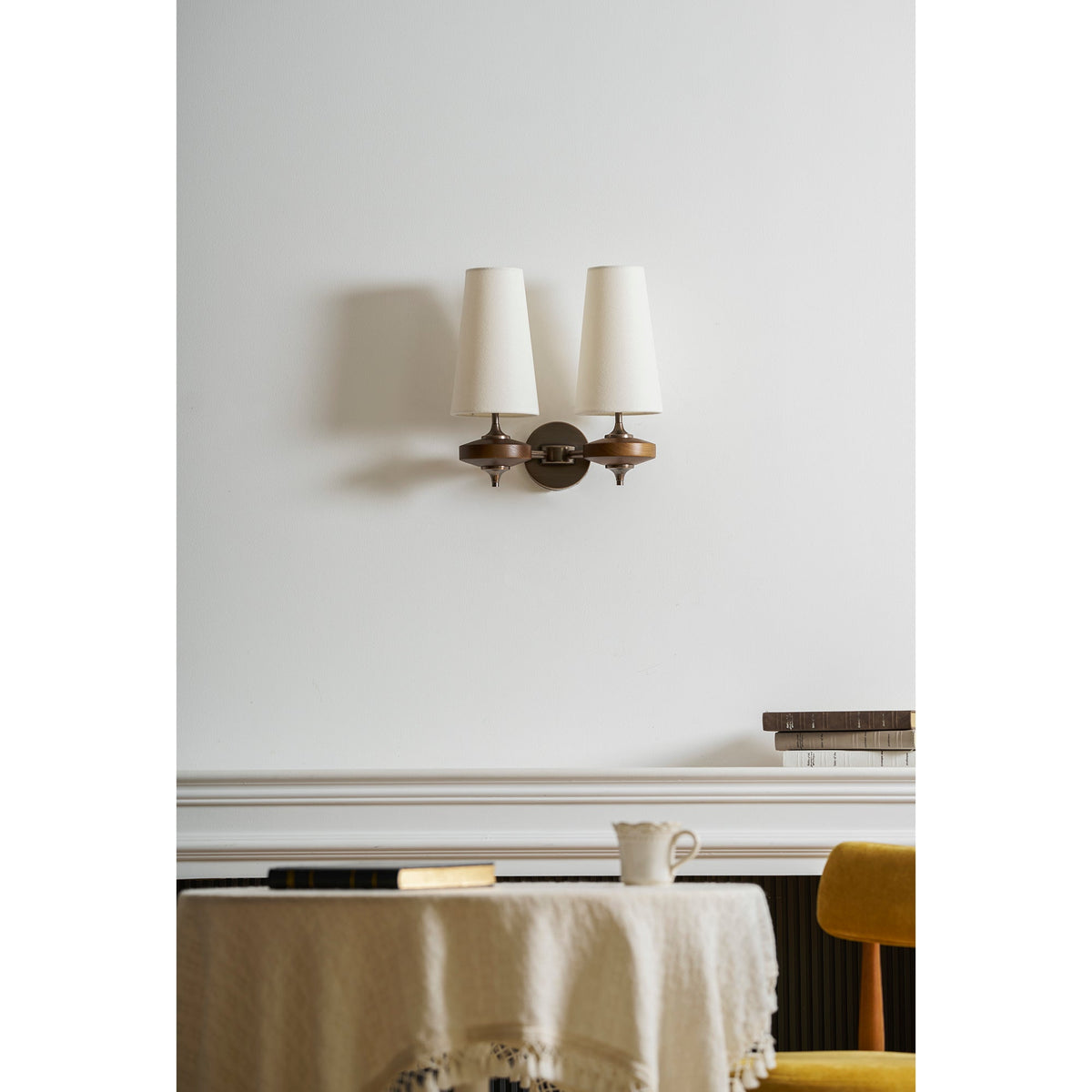 A Clérachic twin wall sconce with cream shades over a white mantel, a small jug and a round table with a linen cloth.