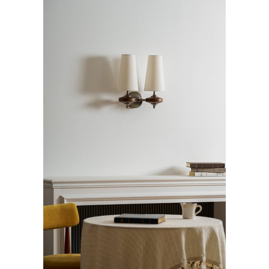 Unlit cream shades on a Clérachic two-light sconce, hung above a white mantel with books and a round linen-covered table.