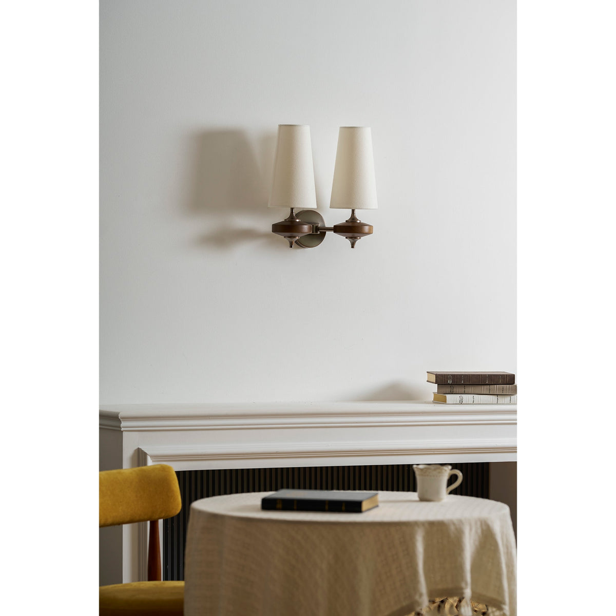 Unlit cream shades on a Clérachic two-light sconce, hung above a white mantel with books and a round linen-covered table.