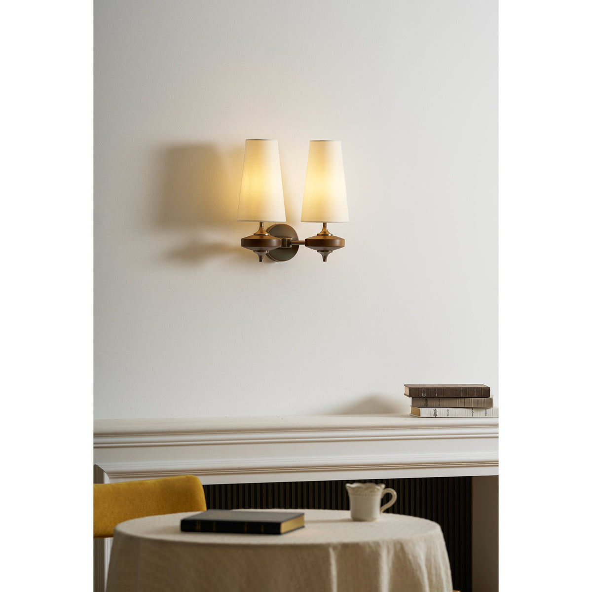 Warm glow from two cream shades on a Clérachic twin sconce, above a mantel, a mustard chair and a laid table.