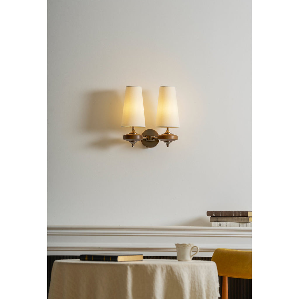 A lit Clérachic twin sconce with cream shades casts a soft pool of light on a white wall above a mantelpiece.