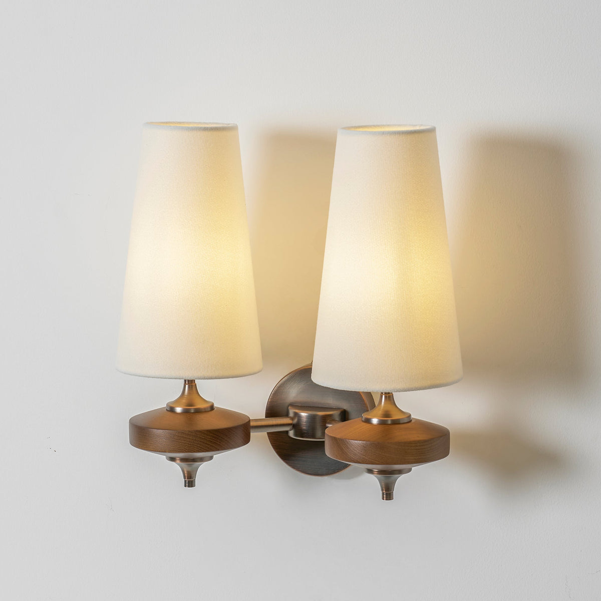 Angled view of a lit Clérachic twin sconce with cream shades, one arm swung forward from the round bronze plate.