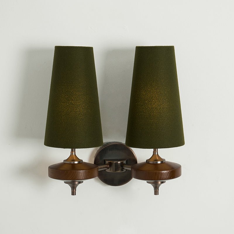 Green shades lit warm from inside on a Clérachic twin wall light, with walnut discs below, on a pale wall.