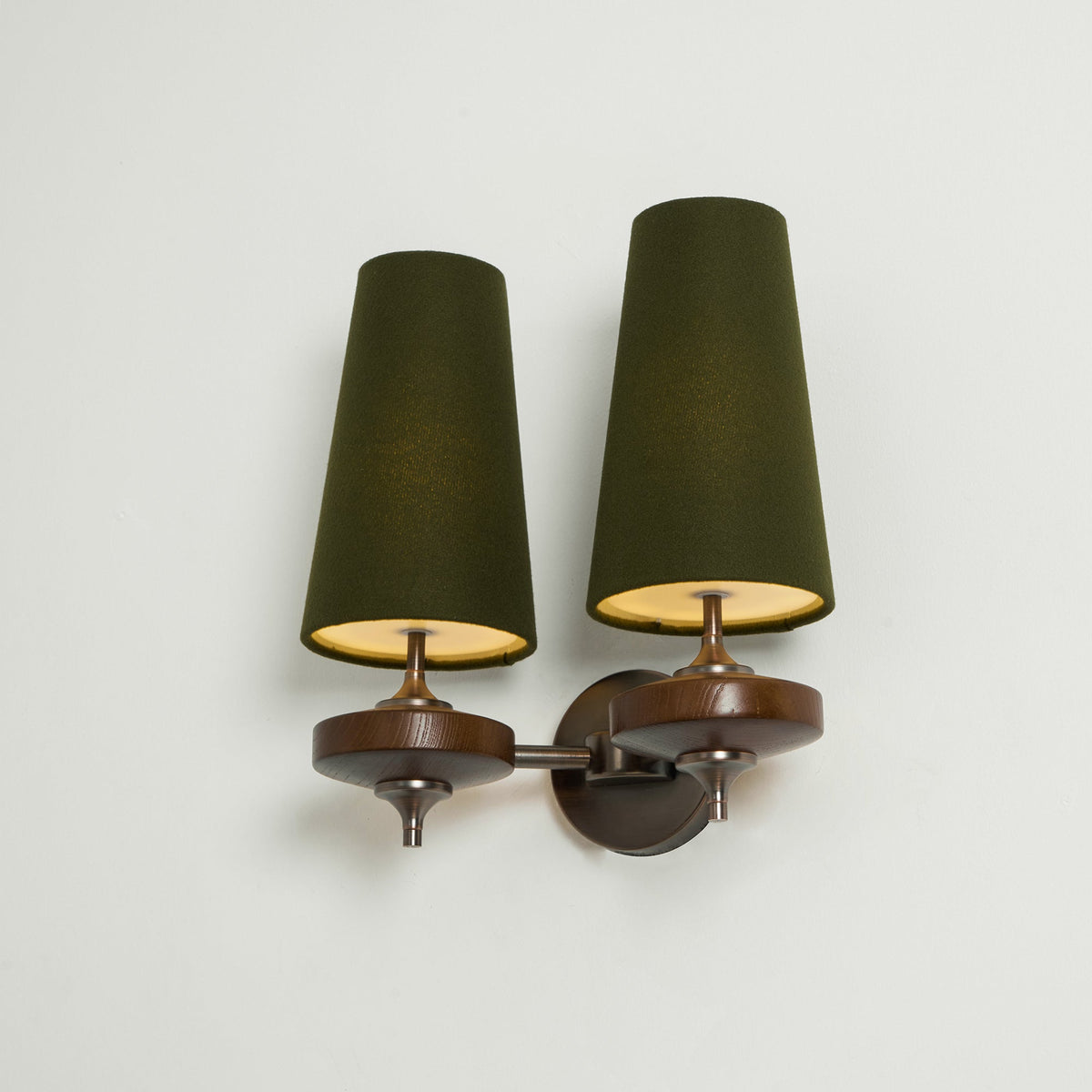 Angled view of a Clérachic twin wall sconce with lit green shades, gold glowing inside, on a pale mint wall.
