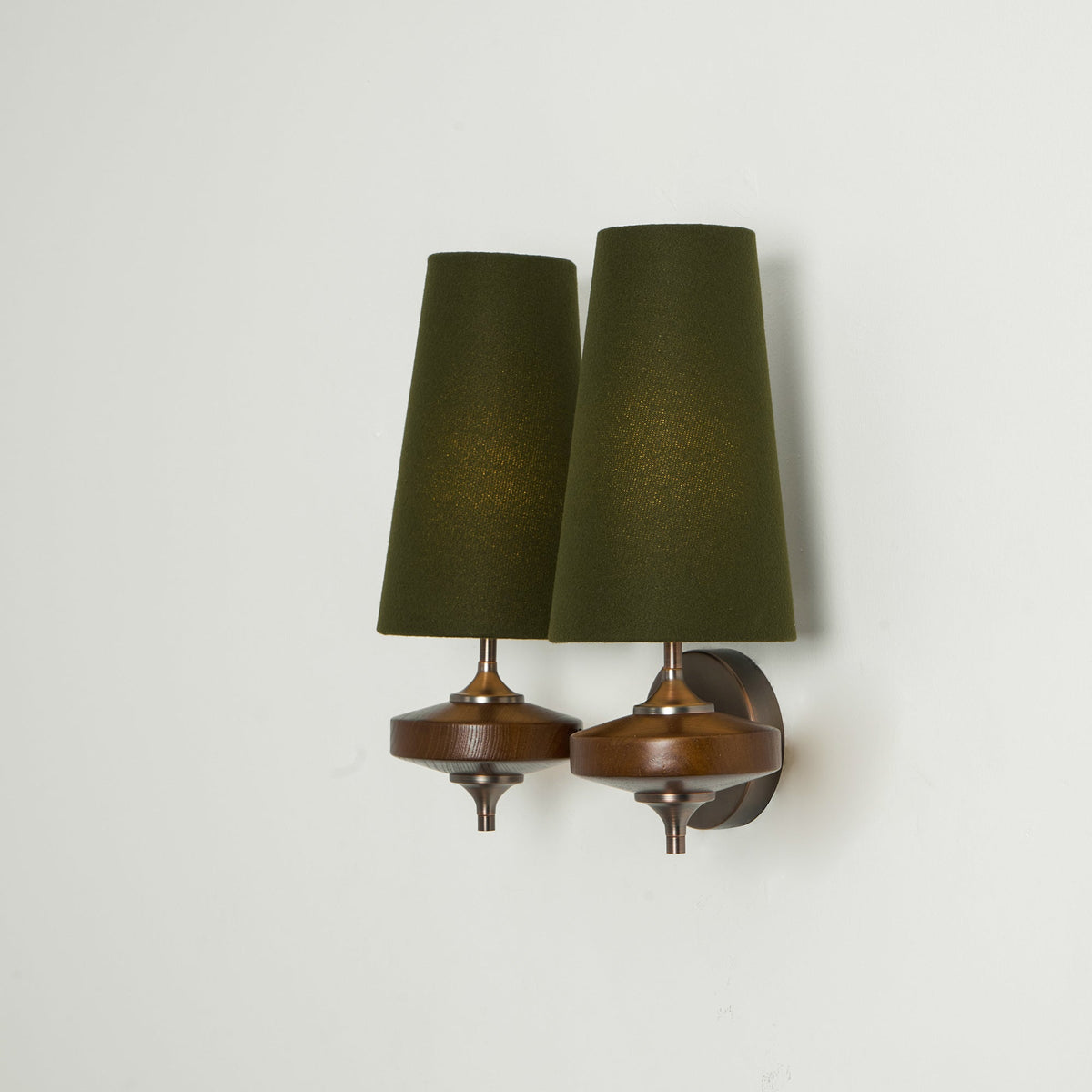 Side-on view of a Clérachic twin sconce with lit green shades and walnut discs, on a pale green-grey wall.
