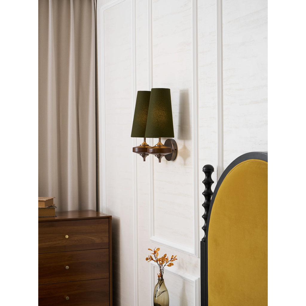A Clérachic green twin sconce seen from the side, beside a taupe curtain, a walnut chest and a mustard headboard.