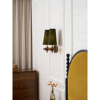 A Clérachic green twin sconce seen from the side, beside a taupe curtain, a walnut chest and a mustard headboard.