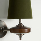 Close view of a green fabric shade, bronze stem and walnut disc on this Clérachic wall sconce, against white.