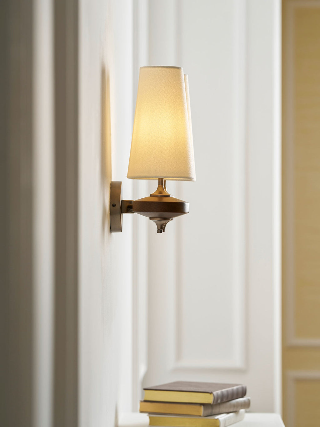 A lit cream shade on a Clérachic wall sconce seen side-on, beside a white panelled door and a stack of books.