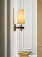 A lit cream shade on a Clérachic wall sconce seen side-on, beside a white panelled door and a stack of books.
