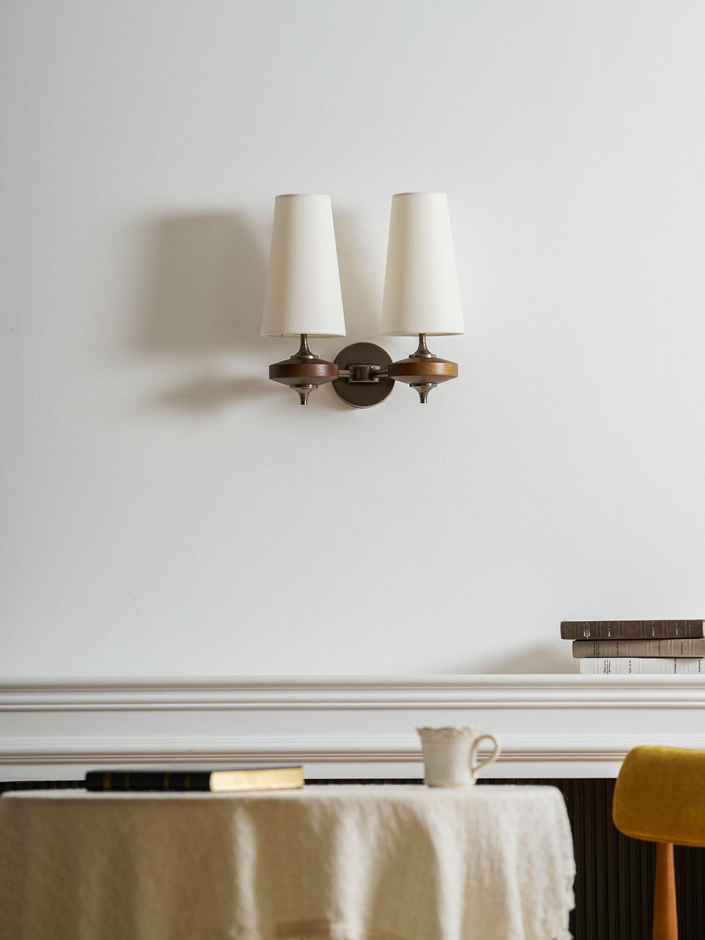 A Clérachic twin sconce with cream shades above a white mantel with books, a jug and a round table with a linen cloth.