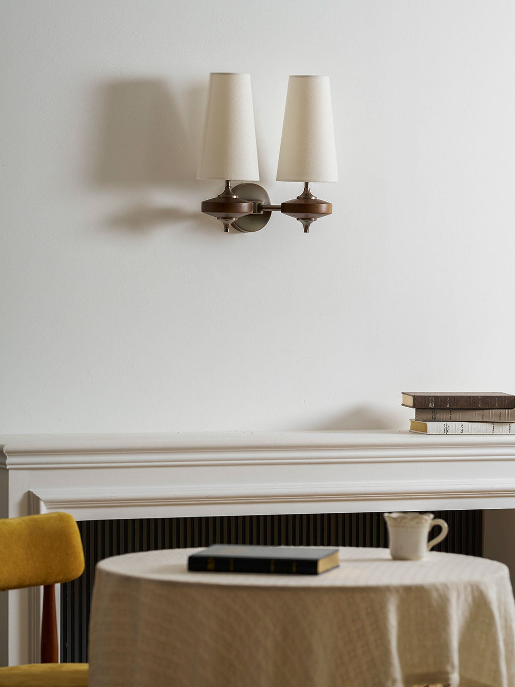 Two unlit cream shades on a Clérachic wall sconce, over a mantelpiece, a round table and a mustard chair.