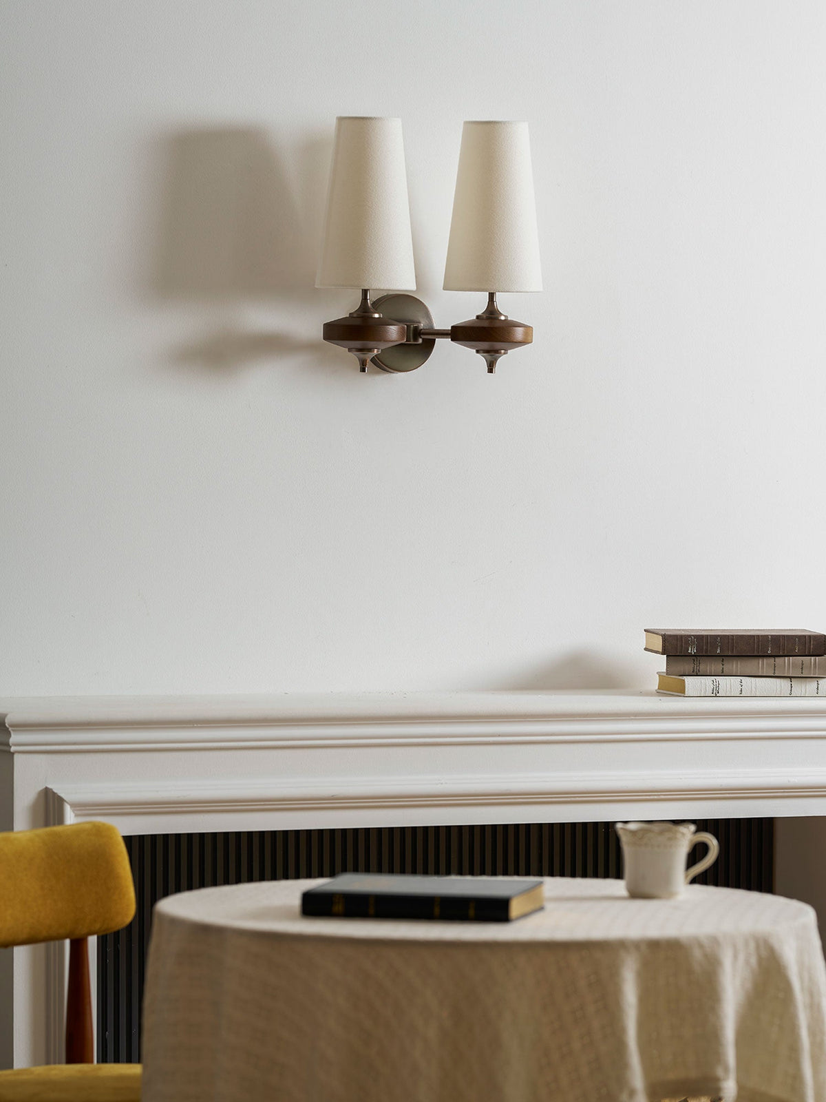 Two unlit cream shades on a Clérachic wall sconce, over a mantelpiece, a round table and a mustard chair.