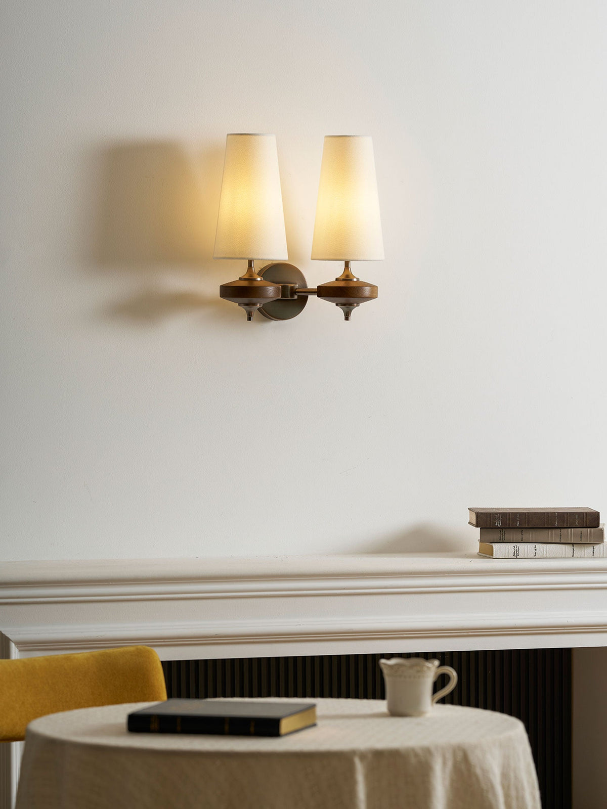 Warmly lit cream shades on a Clérachic twin sconce, above a white mantel with books and a laid round table.