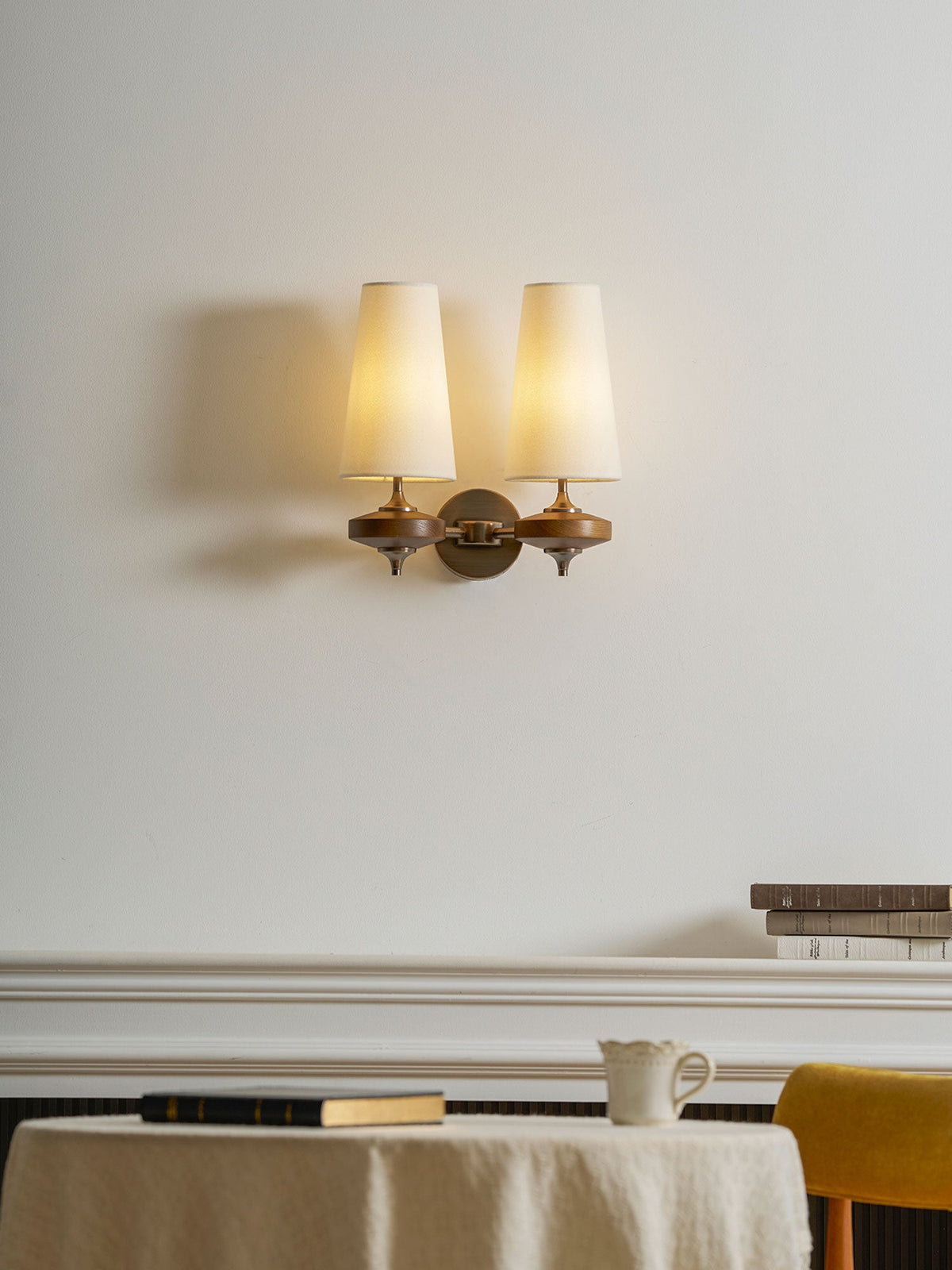 A glowing Clérachic twin sconce with cream shades above a white mantelpiece, a linen-covered table and a mustard chair.