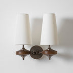 White fabric shades and walnut discs joined by a bronze chain-link arm on a Clérachic twin wall sconce, on white.
