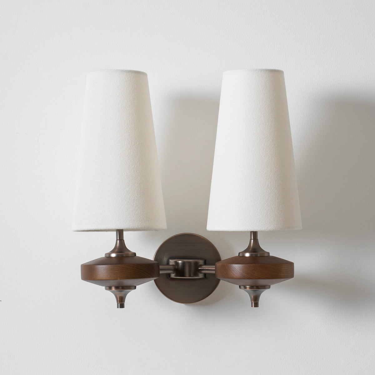 White fabric shades and walnut discs joined by a bronze chain-link arm on a Clérachic twin wall sconce, on white.