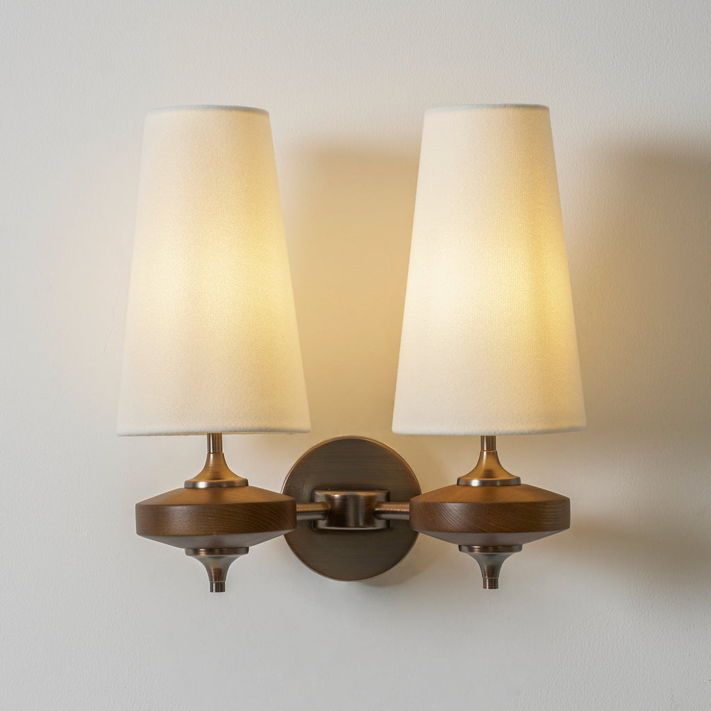 Two lit cream shades fill the frame on this Clérachic twin sconce, with walnut discs and a round bronze backplate.