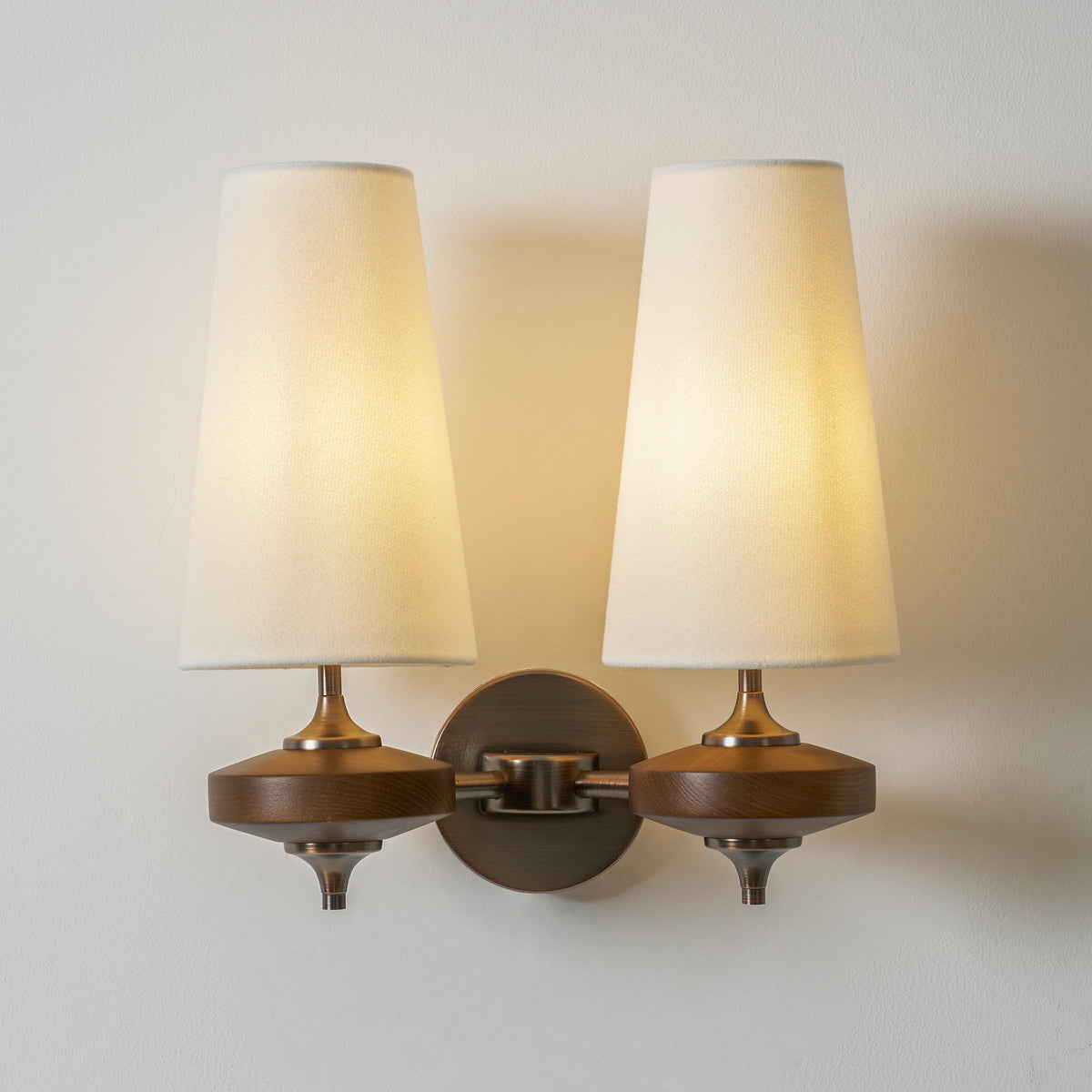 Two lit cream shades fill the frame on this Clérachic twin sconce, with walnut discs and a round bronze backplate.