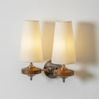 Lit cream shades on a Clérachic twin wall light, one arm swung forward from the bronze backplate, on a pale wall.
