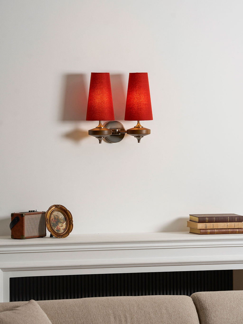 A Clérachic twin sconce with lit red shades above a white mantel, a small retro radio, a round mirror and a sofa.
