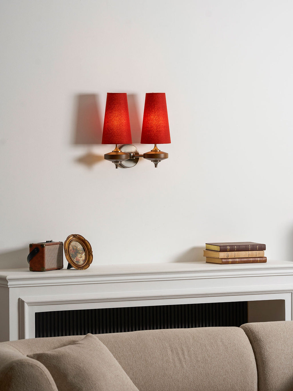 Red shades glow on a Clérachic two-light wall sconce, above a mantel with a vintage box, a gilt frame and a taupe sofa.