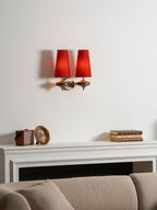 Red shades glow on a Clérachic two-light wall sconce, above a mantel with a vintage box, a gilt frame and a taupe sofa.