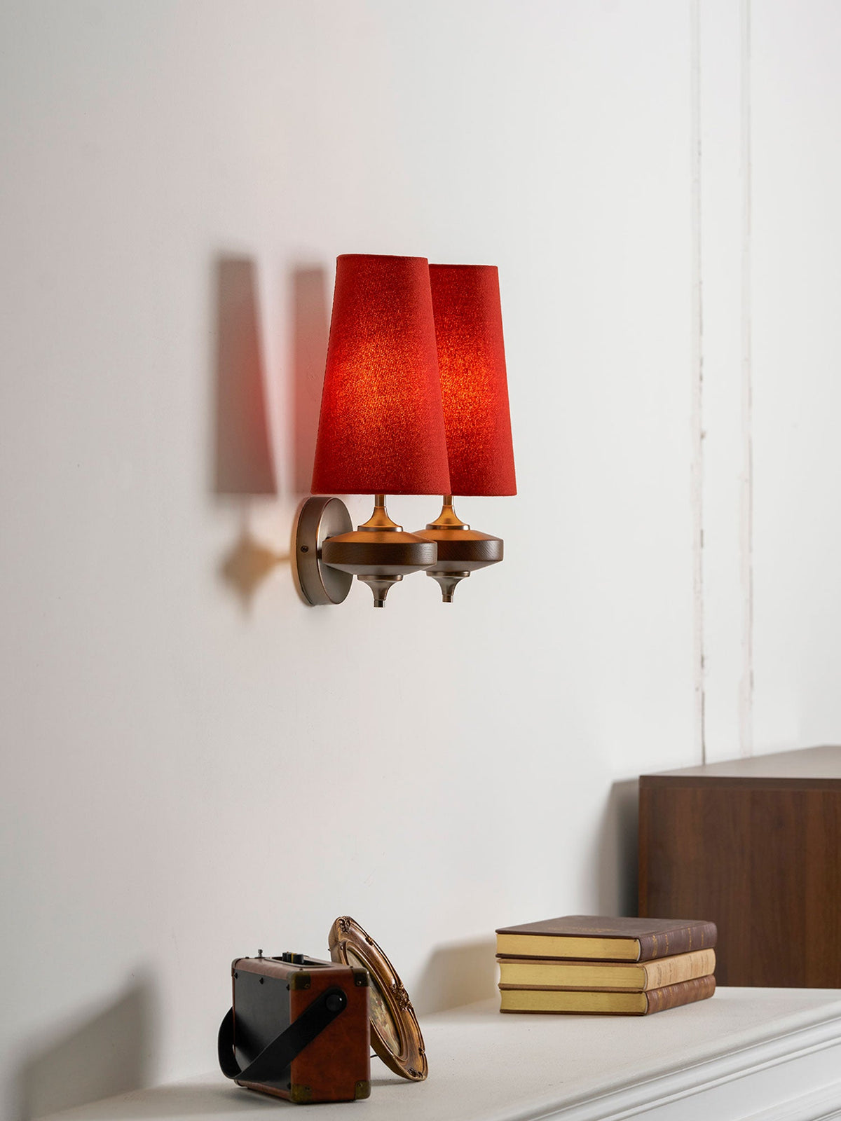 A Clérachic wall sconce with two red shades seen at an angle, above a mantel with a stack of books and an old case.