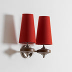 Angled view of a Clérachic twin wall sconce with red shades, one arm turned out, with its shadow cast on a white wall.