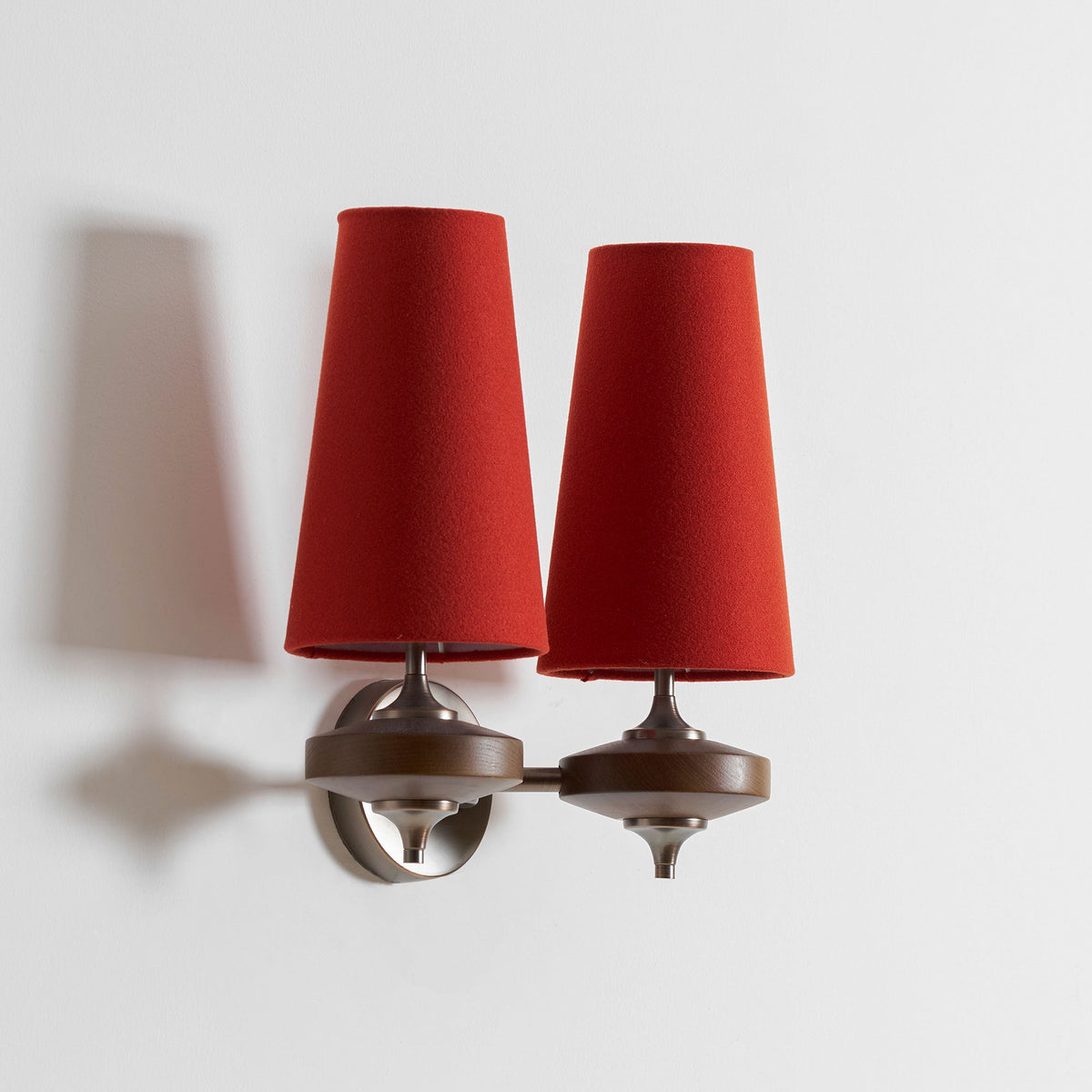 Angled view of a Clérachic twin wall sconce with red shades, one arm turned out, with its shadow cast on a white wall.